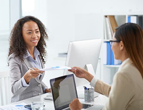 Two professional women exchanging documents at a desk