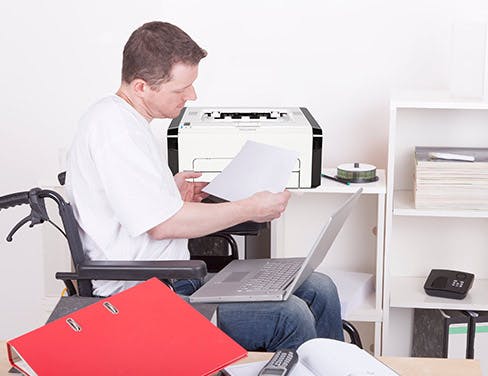 Photo of a man in a wheelchair holding a piece of paper with his laptop in his lap near a printer.