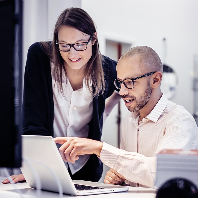 Businessman is sitting in front of a notebook in a modern office room, explaining something to a female colleague standing next to him. He's pointing at the screen while the woman is happily listening to him.