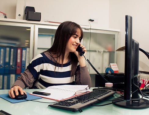 Woman in desk talking on the phone and using desktop computer.