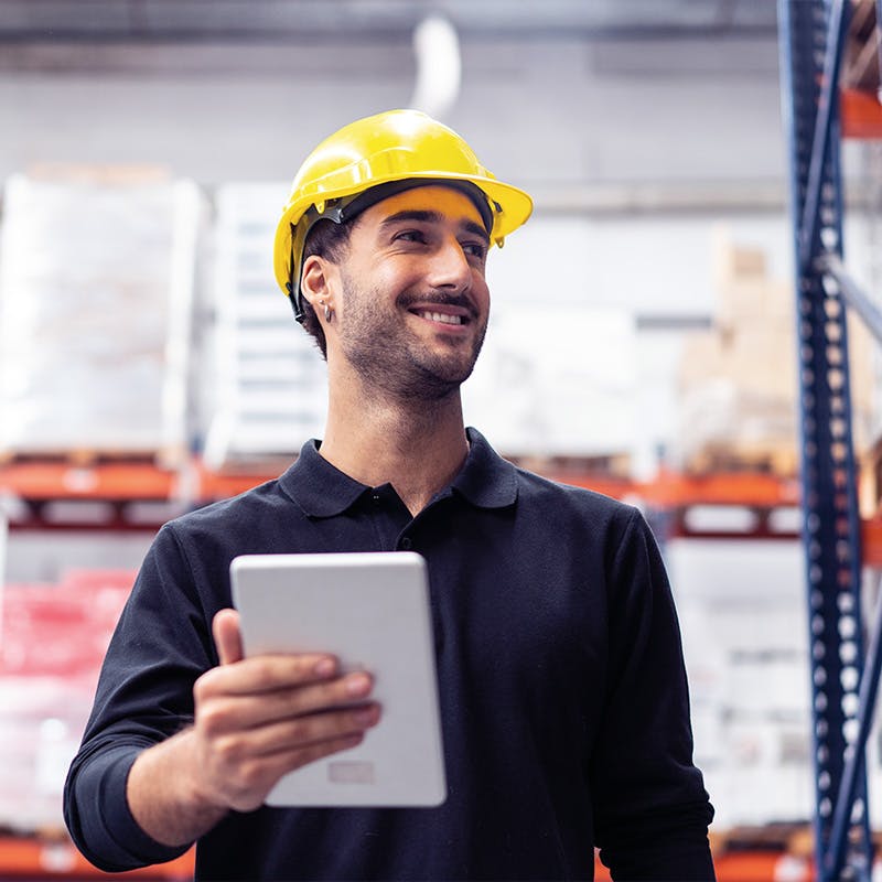 Young man holding a digital tablet in a warehouse. Man working in the warehouse looking away and smiling.
