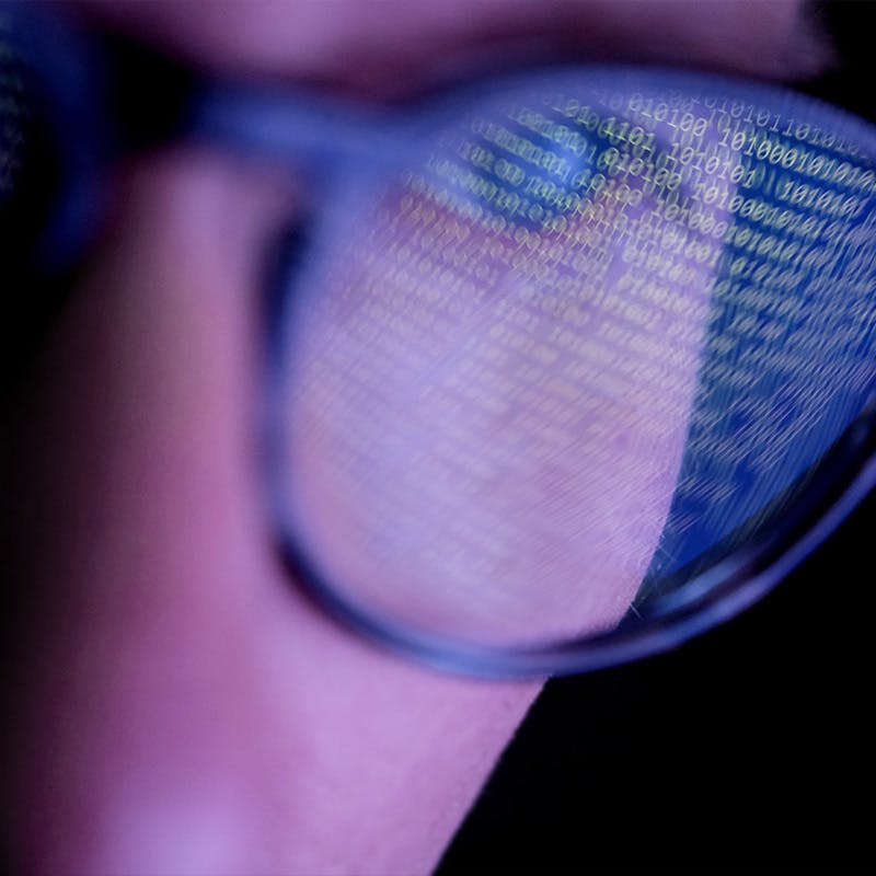 A person with glasses in a dark room looking at computer screen that's displaying codes or programming, with reflections on the glasses