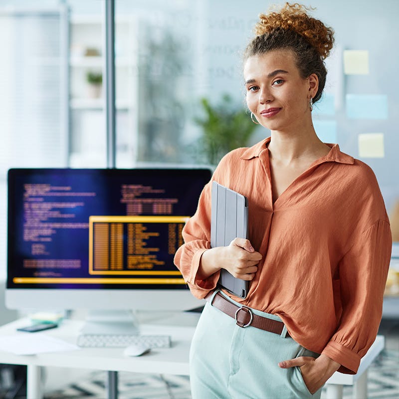 Portrait of young female developer with digital tablet looking at camera while standing at her workplace with computer and coding in the background